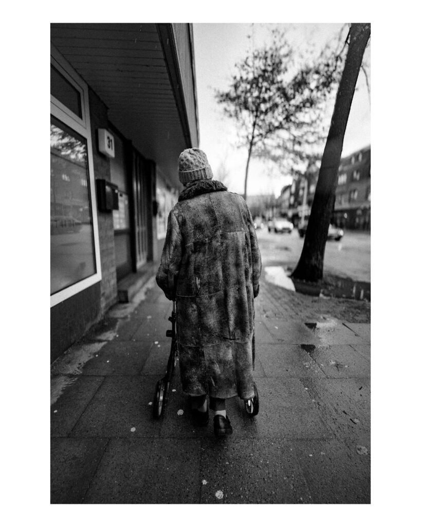 Schwarzweiß-Serie aus Harburg: Eine ältere Frau mit Rollator geht allein über nassen Bürgersteig. Menschen überqueren eine Kreuzung im Gegenlicht.